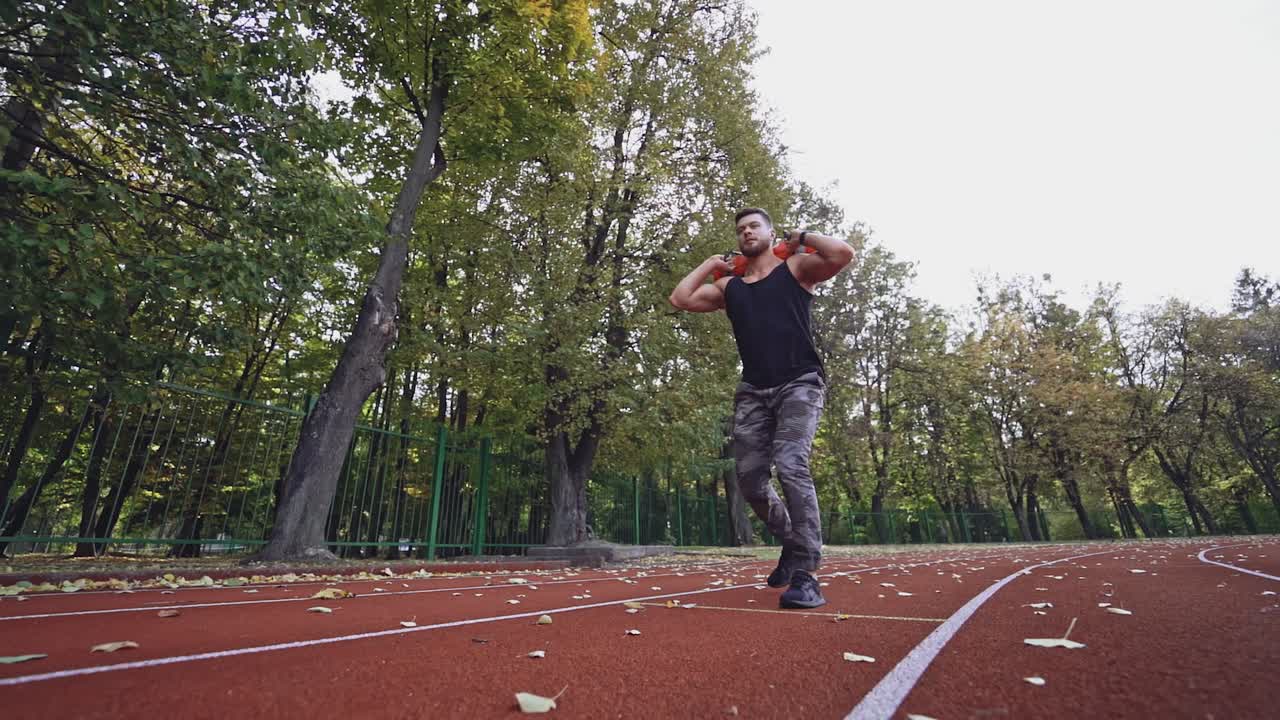 Muscular athlete exercising in the stadium. Young sportsman doing squats with weight while walking. Slow motion.