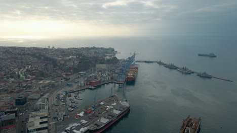 aerial dolly in of valparaiso hillside city and sea port, boats docked and cargo ship near cranes at sunset, chile