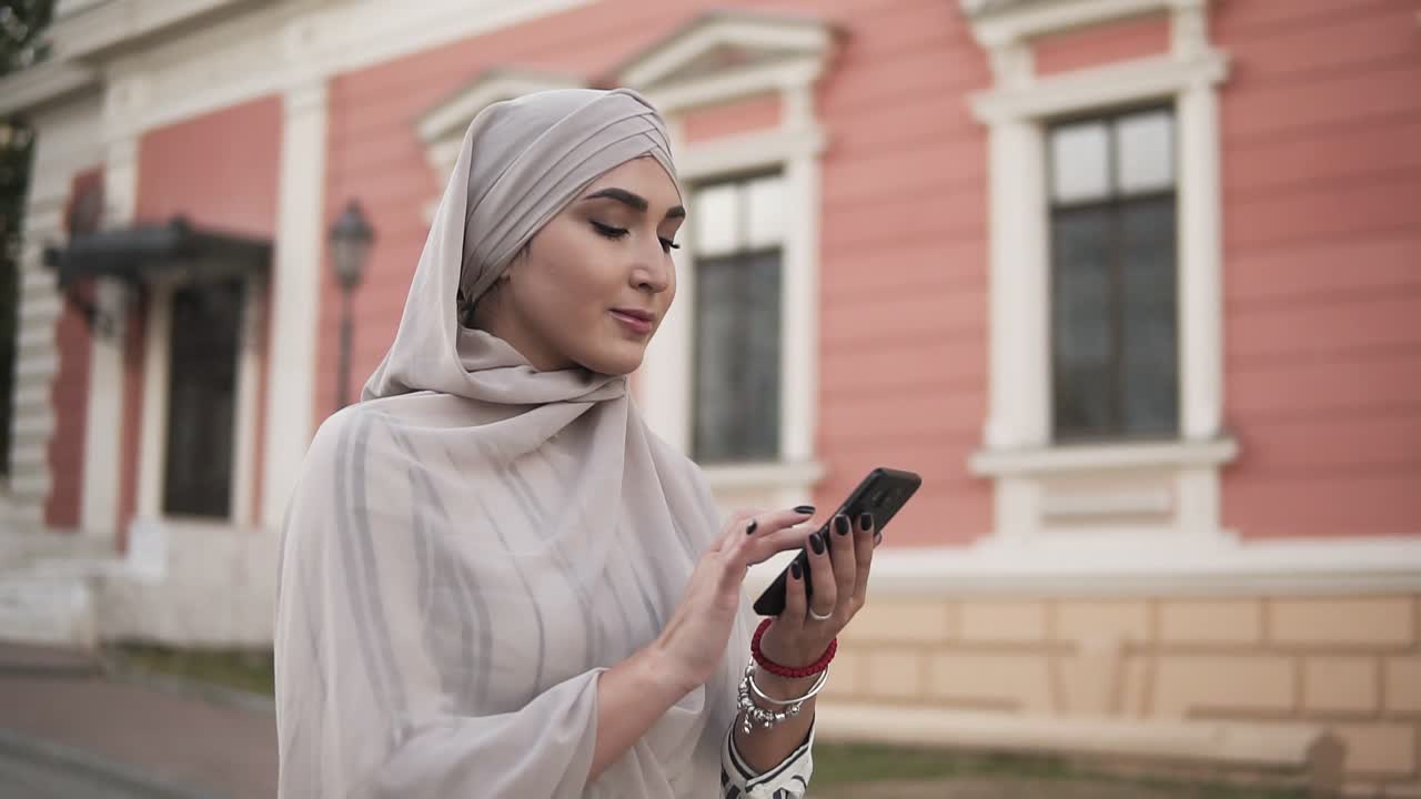 hermosa mujer musulmana en hiyab pastel con maquillaje de pie en la calle sosteniendo un teléfono inteligente mientras camina por la ciudad
