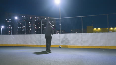 vista trasera de un atleta con capucha pateando una pelota de fútbol contra la pared en un campo deportivo iluminado por la noche, con el reflejo visible en la pared y el paisaje urbano brillando en el fondo