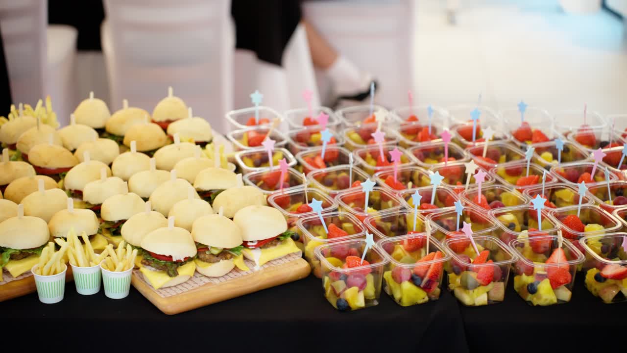A delicious catering spread of mini burger sliders and individual fresh fruit salad cups beautifully arranged for a party or event - A smooth trucking camera shot moves sideways to reveal