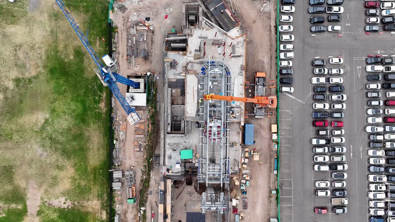 Top-down drone aerial of construction site with orange cranes, steel structure, foundation work, and adjacent organized parking lot