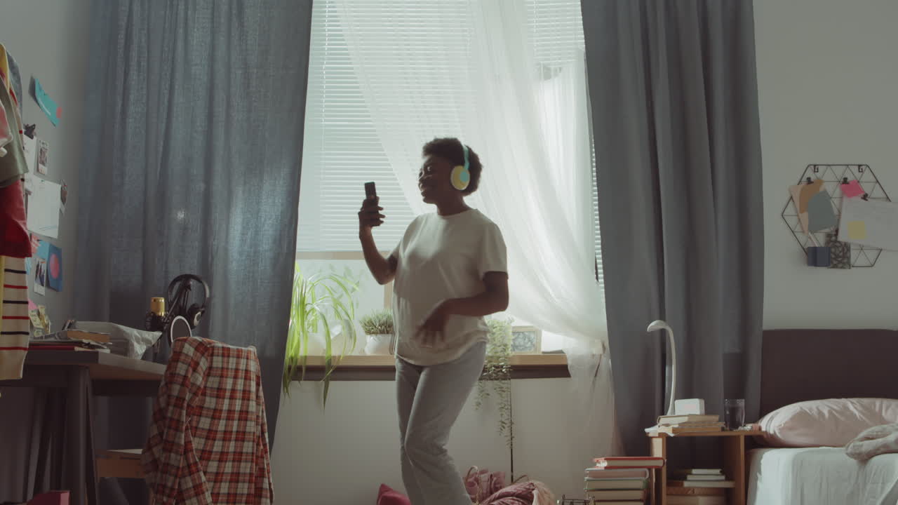 헤드폰을 입은 아프리카계 미국인 여성이 침실에서 춤을 추고 있습니다.