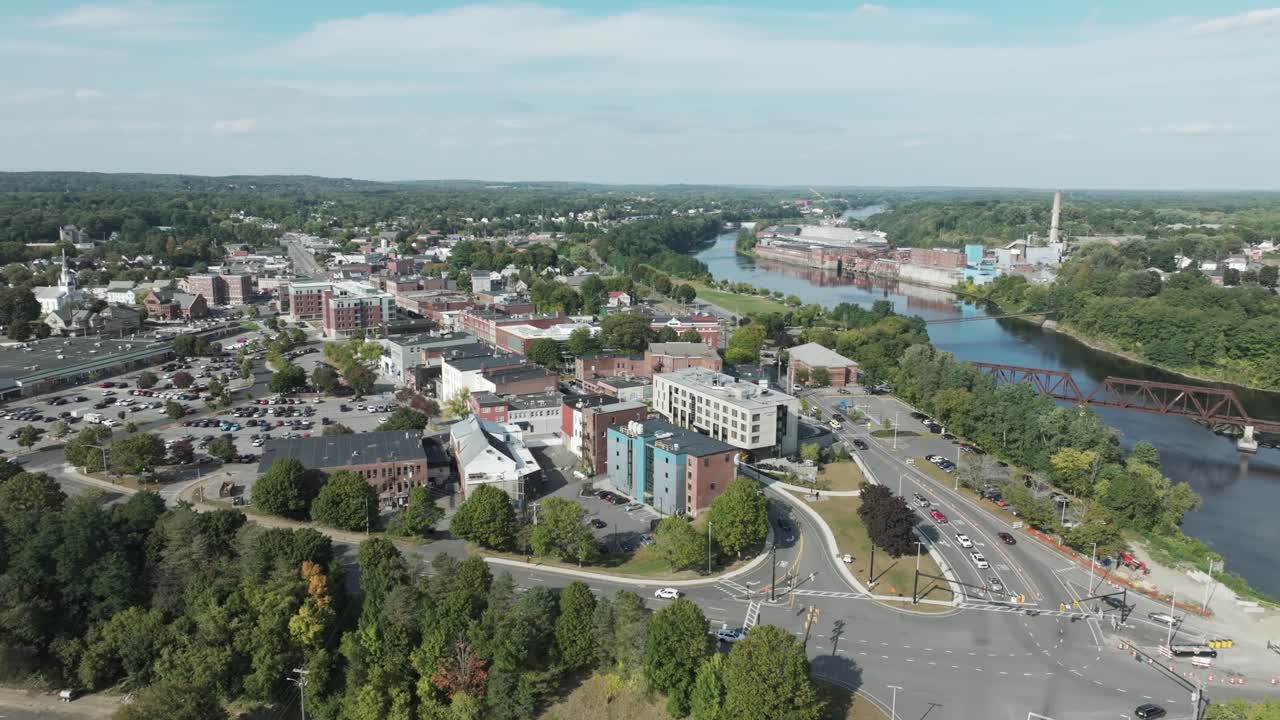 Aerial view of downtown Waterville, Maine showing city streets, modern buildings, the Kennebec River, and a historic railway bridge on a clear summer day.