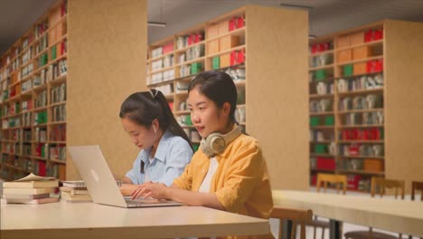 asian woman student with headphones typing on a laptop while sitting with her classmate studying on a table in the library