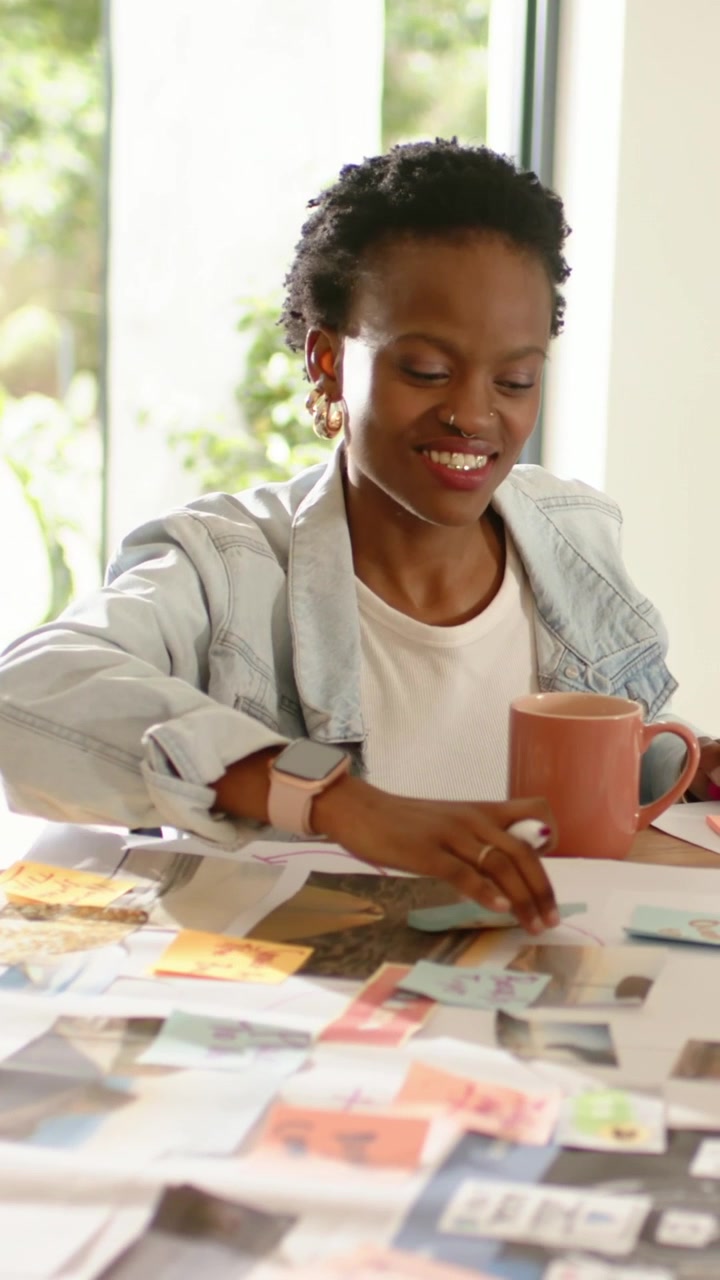 Vertical video: Pen twirling, woman in denim sorting notes and steadying mug in studio for layout