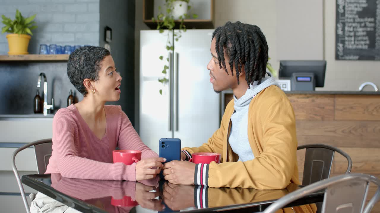 Diverse couple leaning at glass kitchen table noticing smartphone screen chatting over coffee
