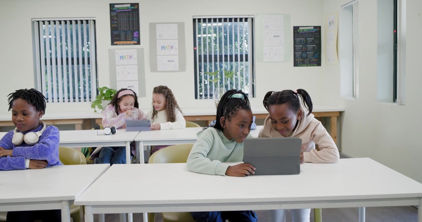 Diverse female students focusing on tablets, shifting closer and sharing tablet at school desks