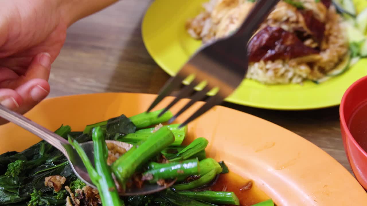 Hands use fork and spoon to transfer stir-fried Chinese broccoli with sauce from orange plate to yellow plate of roasted rice in bright, casual dining setting