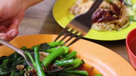 hands use fork and spoon to transfer stir-fried chinese broccoli with sauce from orange plate to yellow plate of roasted rice in bright, casual dining setting