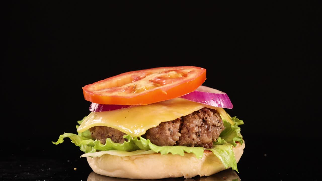 A sesame seed bun top falls onto a beef burger with lettuce, tomato, onion, and cheese, shot in slow motion against a black background with studio lighting