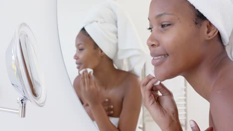 Happy-biracial-woman-with-towel-on-head-looking-in-mirror-washing-her-face,-slow-motion