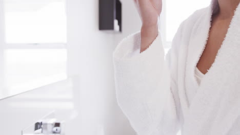 Biracial-woman-applying-cleansing-face-using-cotton-pad-looking-in-mirror-in-bathroom,-slow-motion