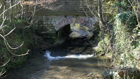 4K-Aufnahmen-Eines-Flusses,-Der-Unter-Einer-Kleinen-Steinbrücke-Hindurchfließt