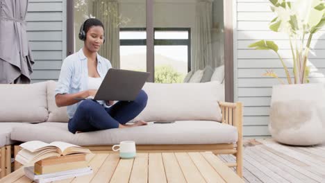 african american woman sitting on sofa on terrace wearing headphones, using laptop, slow motion