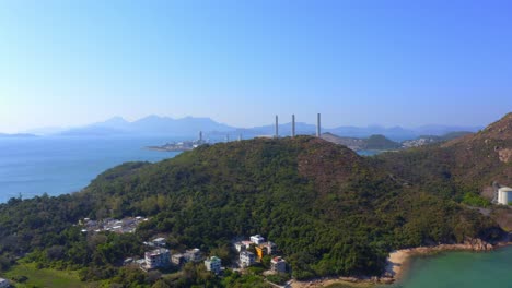 아름다운 화창한 날 해변이 있는 마을 옆에 있는 섬에 있는 큰 해안 석탄 화력 발전소를 드러내기 위해 큰 녹색 산으로 앞으로 나아가는 드론 샷