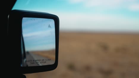 handheld footage of car driving on american west highway, showcasing flat fields and blue skies in car's side mirror