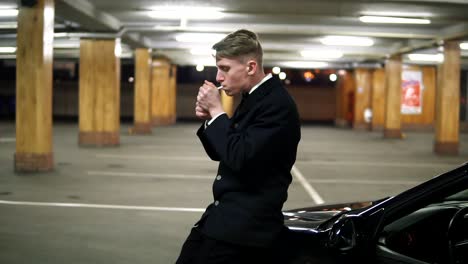 Un-Joven-Con-Traje-Negro-Y-Pajarita-Fumando-Un-Cigarrillo-Sentado-En-El-Capó-Del-Auto-Negro-En-El-Estacionamiento.-Esperando