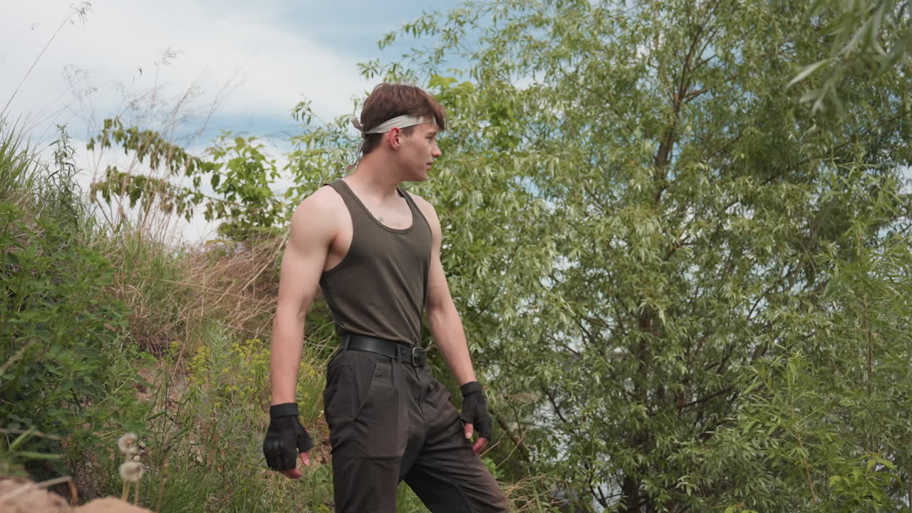 military man jumping down steep riverbank under bright cloudy sky framed by riverside trees in tactical attire demonstrating dynamic movement and adventurous spirit in outdoor environment