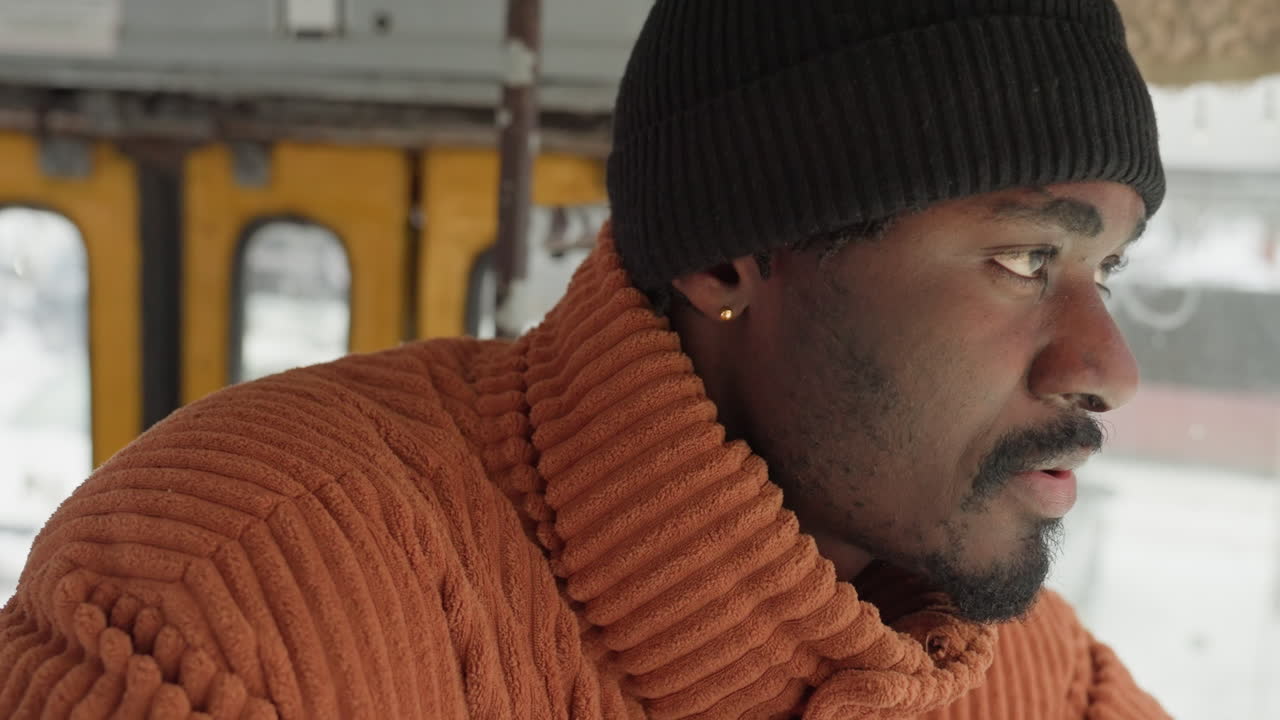 Portrait rapproché de profil d'un homme noir, conducteur de tram contemplatif regardant à travers une fenêtre enneigée, pull en maille orange et bonnet noir, détail de boucle d'oreille, col en laine texturée, lumière naturelle douce, transport urbain