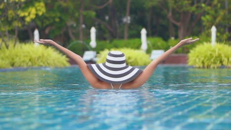 Vista-Trasera-De-Una-Turista-De-Pie-Medio-Sumergida-En-La-Piscina-Con-Un-Gran-Sombrero-Blanco-Y-Negro,-Levantando-Los-Brazos-Por-Encima-De-La-Cabeza,-Disfrutando-De-Sus-Vacaciones-Tropicales