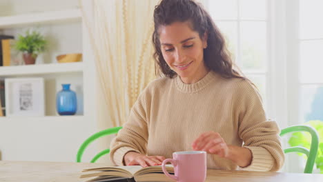 Mujer-Relajándose-En-Casa-Sentada-En-La-Mesa-Leyendo-Un-Libro-Con-Una-Bebida-Caliente