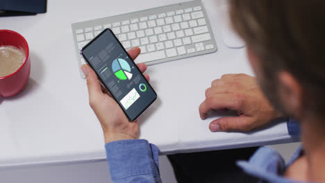 hombre caucásico sentado en un escritorio usando un teléfono inteligente con estadísticas en la pantalla