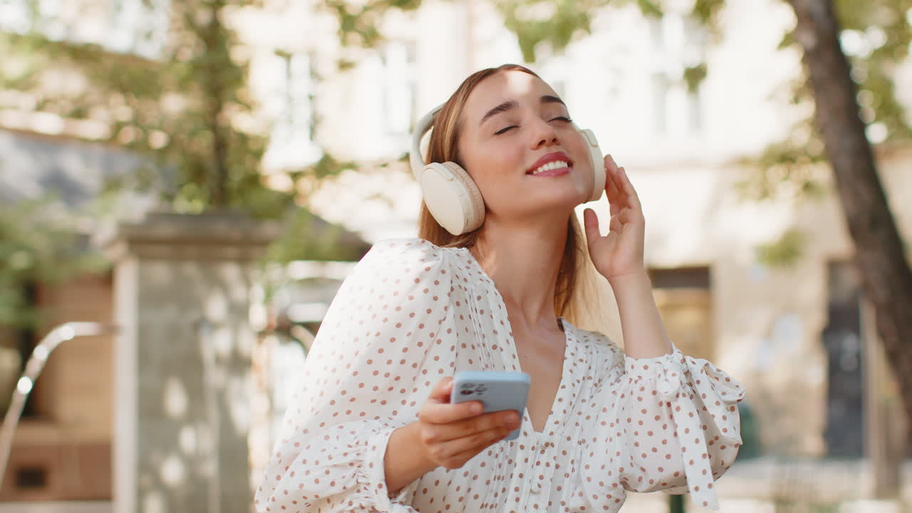 Happy relaxed woman standing on city street in wireless headphones listening dancing music outdoors