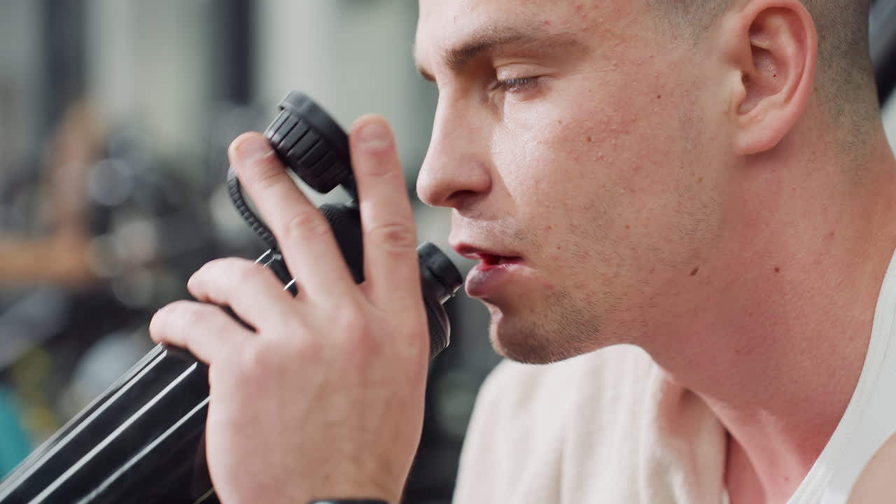 Close up of trainer drinking water with relief on face during rest on weight bench after intense chest press session in modern gym setting under bright lights, recovering strength and rehydrating
