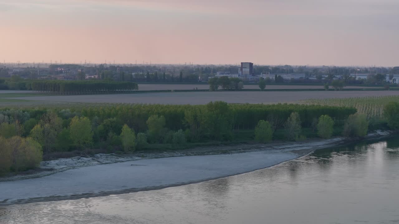 Slow drone pan captures the Po River beach at sunset with golden light over riparian trees, alluvial fields, and Cremona’s skyline showing layered terrain and natural-urban transition