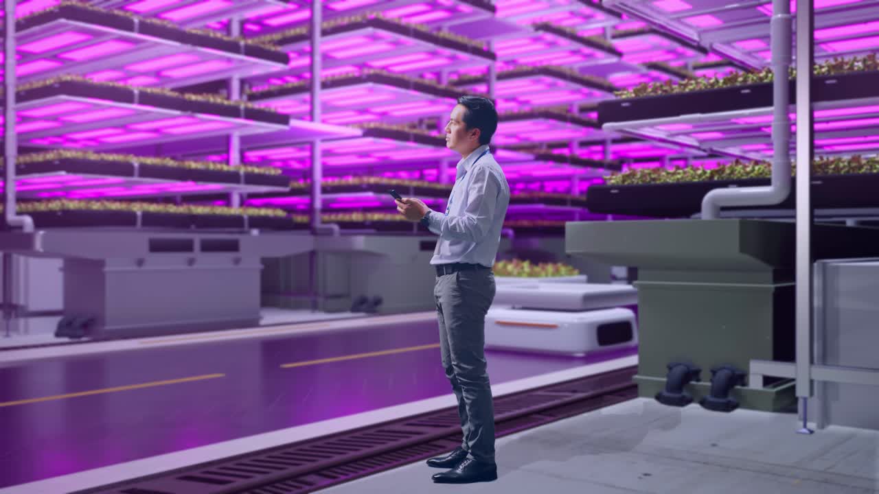 vista lateral del cuerpo completo de un trabajador masculino asiático de pie con su teléfono en una granja vertical, instalación industrial de alta tecnología, observa mirando a su alrededor antes de concentrarse con su teléfono inteligente