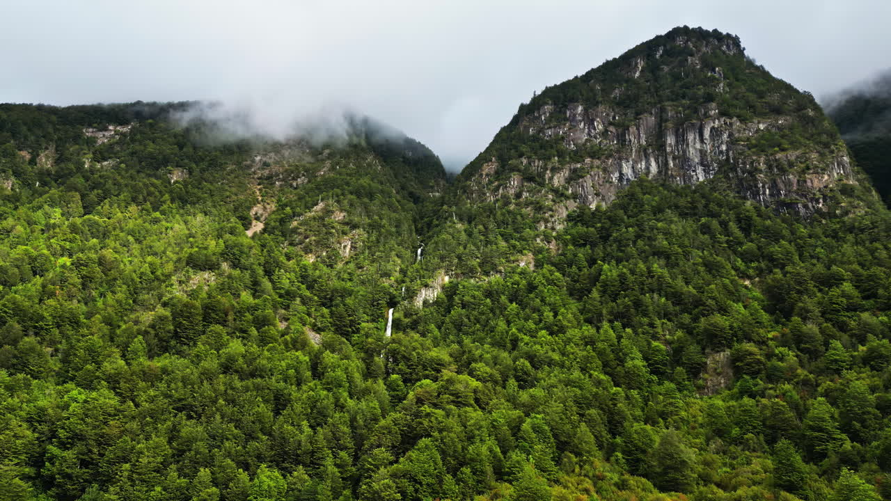 Aerial drone view of a tall waterfall cascading through dense green forest between misty mountains in Patagonia