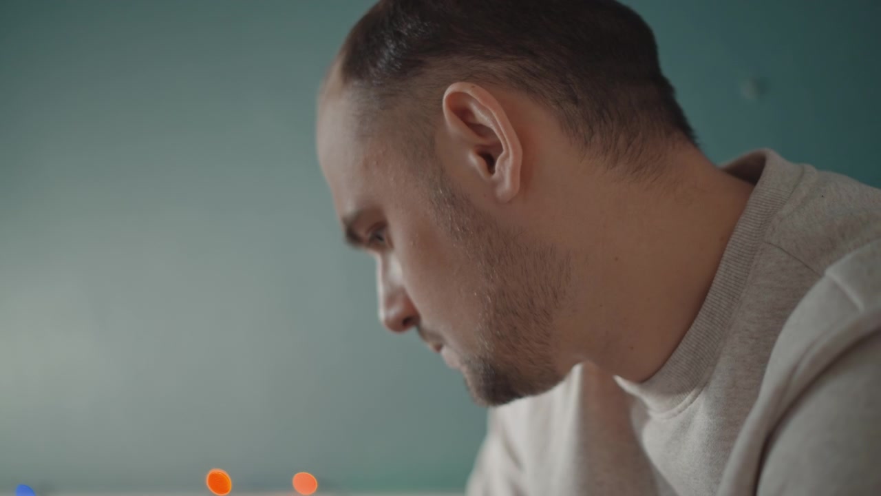 Adult man in three-quarter profile tilts head downward with calm focused gaze and slight forward lean, creating quiet indoor portrait mood with thoughtful concentration, natural stillness and subtle
