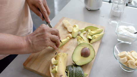 midsection of senior biracial man chopping fruit and vegetables for smoothie in kitchen, slow motion