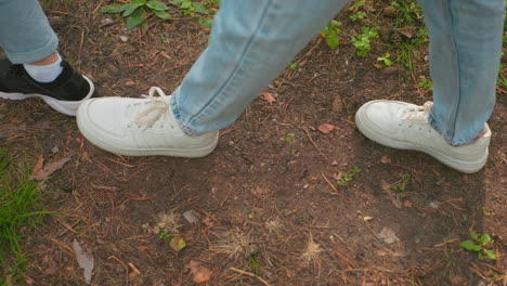 primer plano de dos individuos caminando por el sendero del bosque en vaqueros azules, uno con zapatillas negras y el otro con zapatillas blancas, la escena captura el suelo del bosque de tierra cubierto de agujas de pino, hojas esparcidas