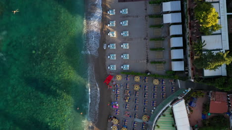 aerial top down view of a sand beach with colorful sunbeds organized into rows. a bird's eye view of clear mediterranean sea with foaming waves, a few people swimming. city of taormina, sicily, italy