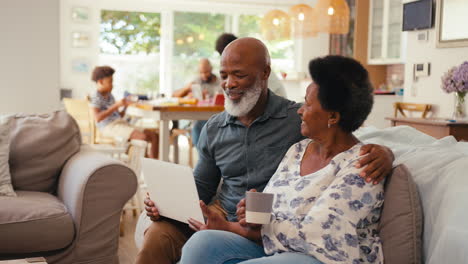 Senior-Couple-At-Home-Looking-At-Laptop-Together-With-Multi-Generation-Family-In-Background