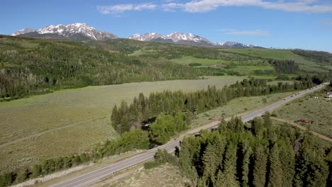 suv blanco conduciendo a lo largo de la carretera de montaña entre los árboles durante el día con montañas cubiertas de nieve, aérea