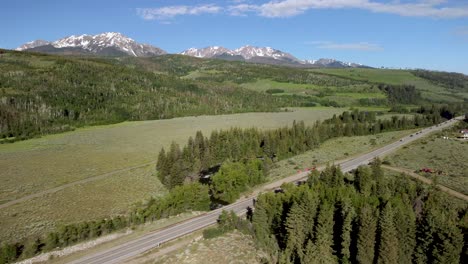 white suv driving along mountain road between trees during the day with snowcapped mountains, aerial