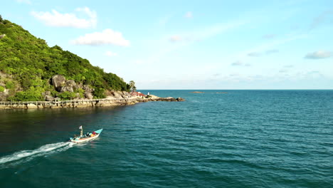 Drone-Siguiendo-Un-Barco-Con-Playa-Y-Montañas-En-El-Fondo