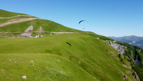 lone paraglider flying across a lush green hill with mountain peaks in the background