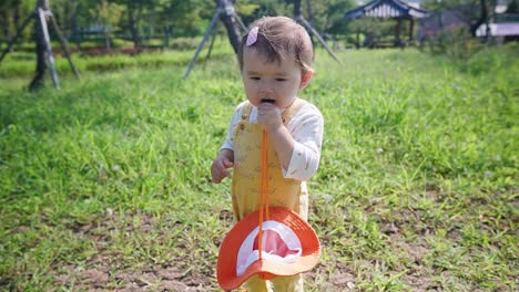 cute asian-caucasian multi-ethnic baby in a city park standing in the grass and holding her hat