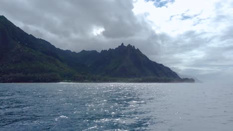 4K-Hawaii-Kauai-Boating-on-ocean-floating-right-to-left-with-left-to-right-pan-of-backlit-mountains-along-cloudy-shoreline-with-ocean-spray-in-foreground