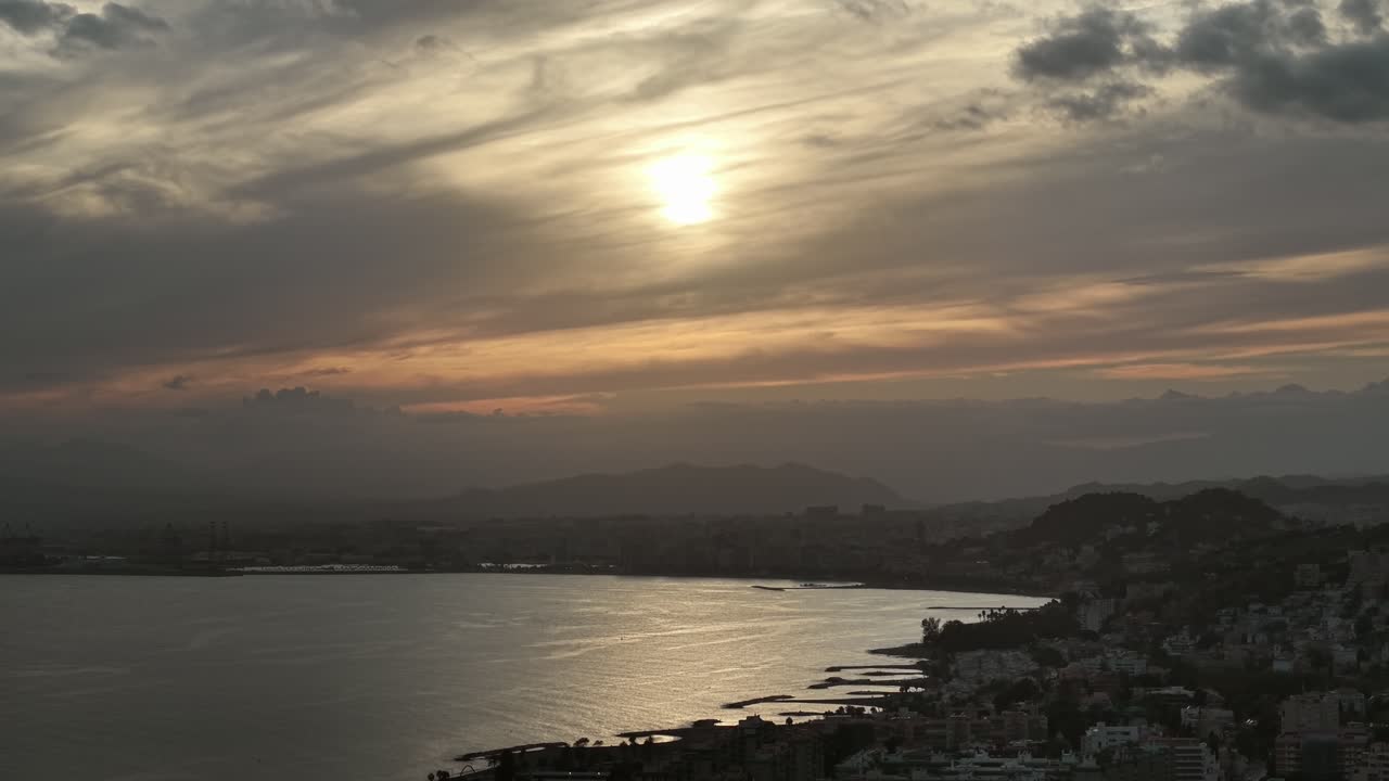 Aerial view of the Málaga coast under a dramatic cloudy sunset, Andalusia, Spain. The golden light reflects on the calm sea and city skyline, evoking a serene yet cinematic Mediterranean atmosphere