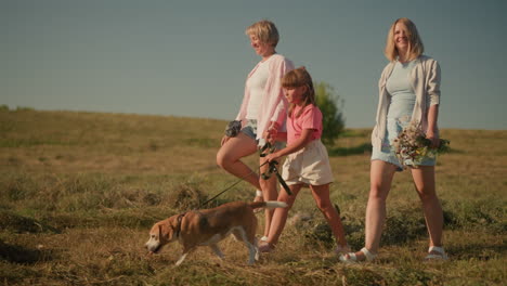 two women and young girl walking in grassy field, girl holding dog leash while dog pauses to look up at one woman, woman in blue carries flowers, capturing joyful moment of outdoor adventure