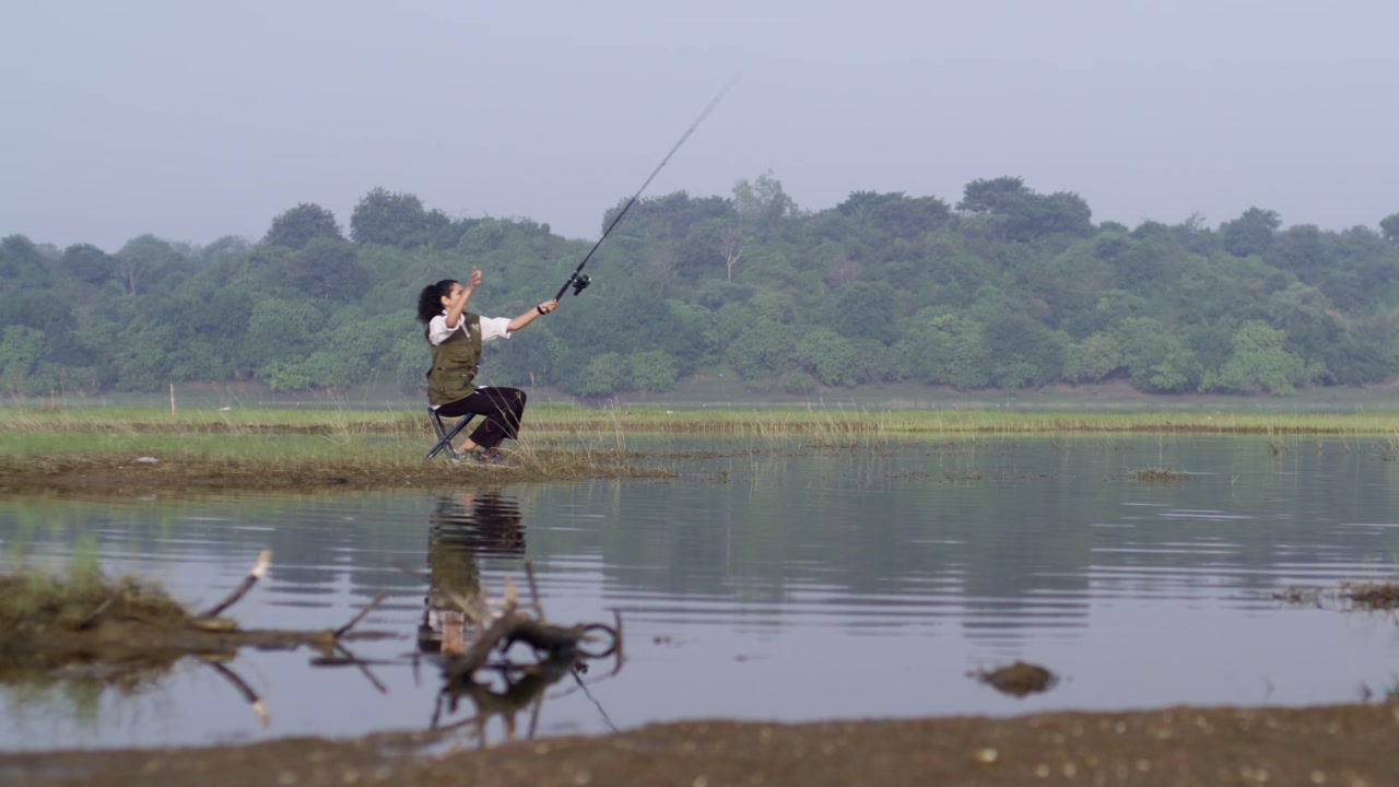 A South Asian woman casting a fishing rod while sitting on a chair by a calm lakeside, enjoying rural nature, water reflections, and peaceful daylight