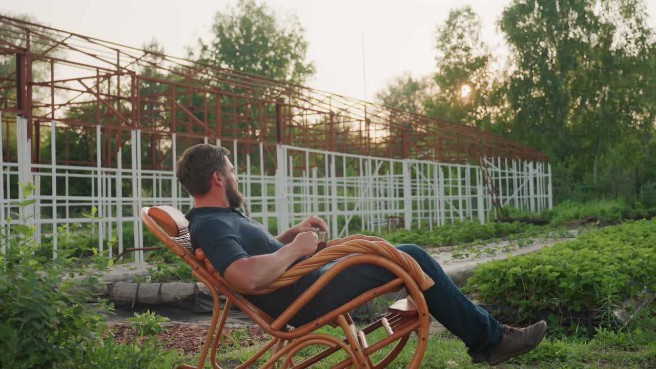 gardener seated on wicker rocker chair leaning back gazing into distance holding clear mug of steaming coffee metal frame and lush garden plants behind under warm sunset light