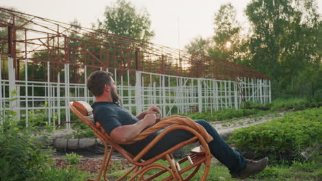 gardener seated on wicker rocker chair leaning back gazing into distance holding clear mug of steaming coffee metal frame and lush garden plants behind under warm sunset light