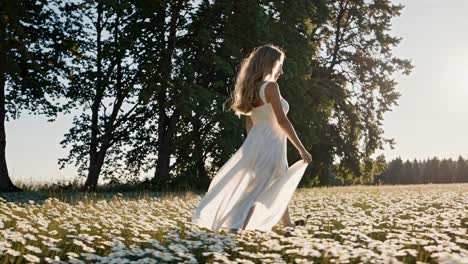 A woman in a flowing white dress walks through a daisy field at sunset