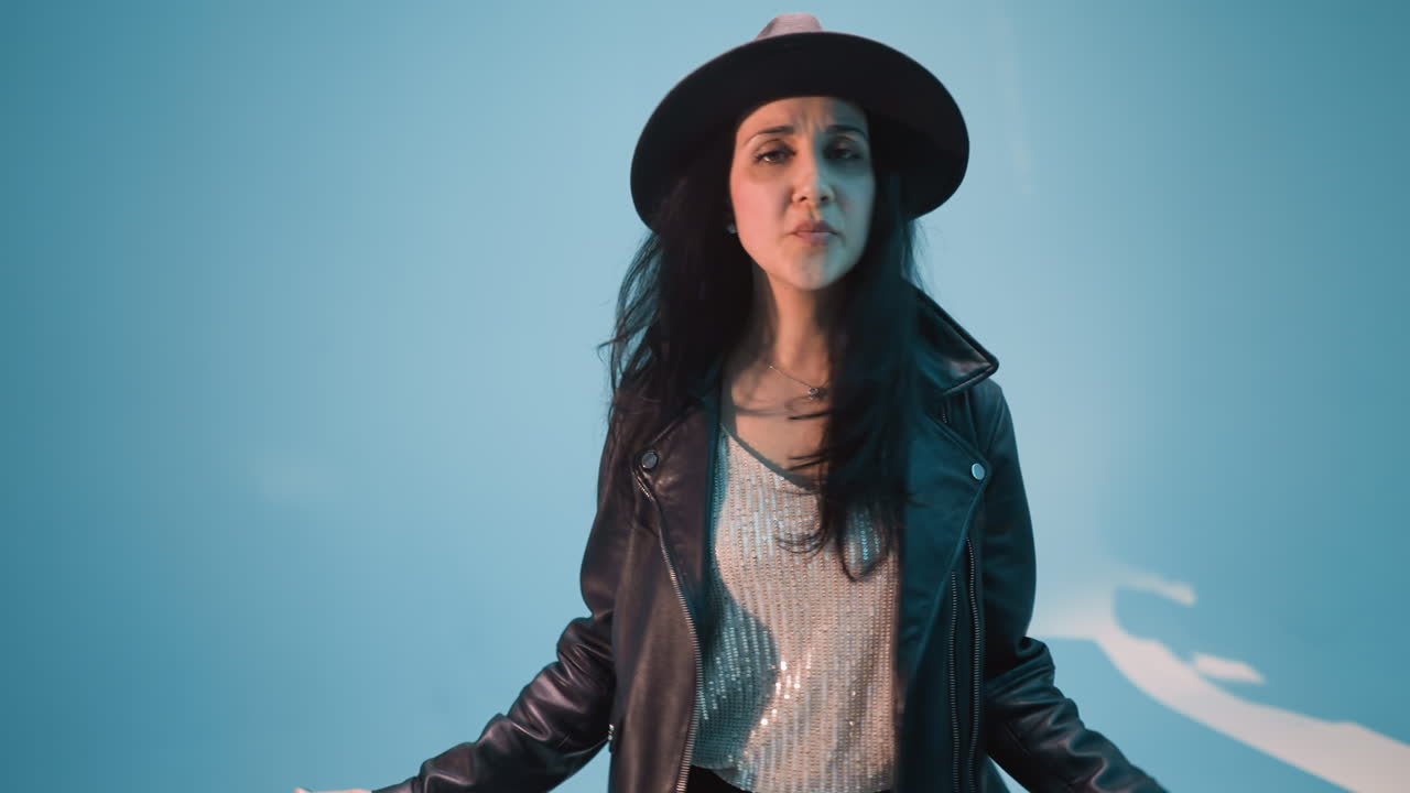 una mujer con una chaqueta de cuero negra, pantalones y sombrero se para y canta sin micrófono. mueve su cuerpo y manos lentamente contra un fondo azul